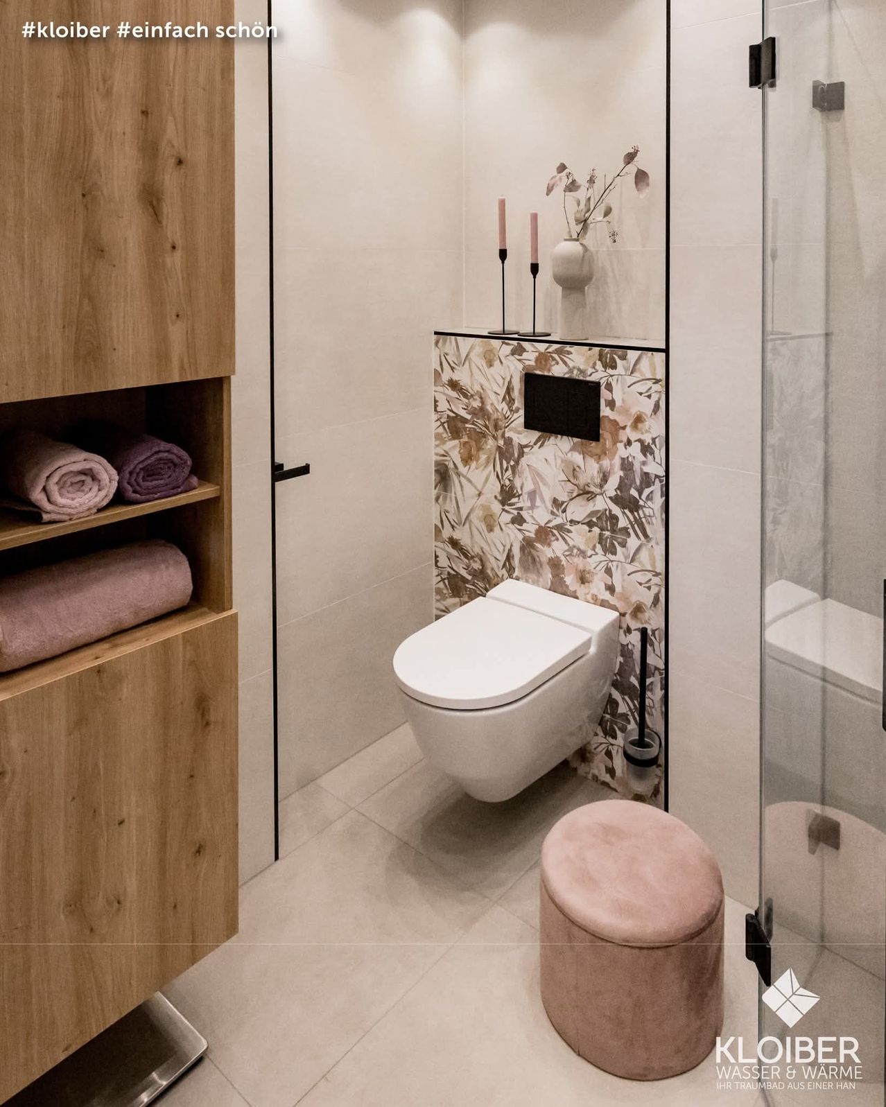 Ein modernes Badezimmer mit weißem WC, floralem Tapetenmuster und einem Holzschrank mit gefalteten Handtüchern. Zwei Kerzen und eine Vase mit Blumen verleihen einen Hauch von Eleganz.
