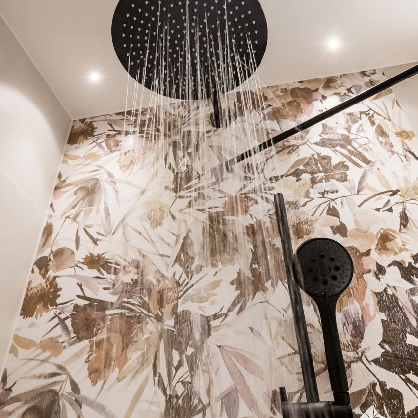 Eine moderne Dusche mit schwarzem Duschkopf und blumengemusterter Wand. Wasser fließt aus dem Duschkopf und bildet einen sanften Nebel.