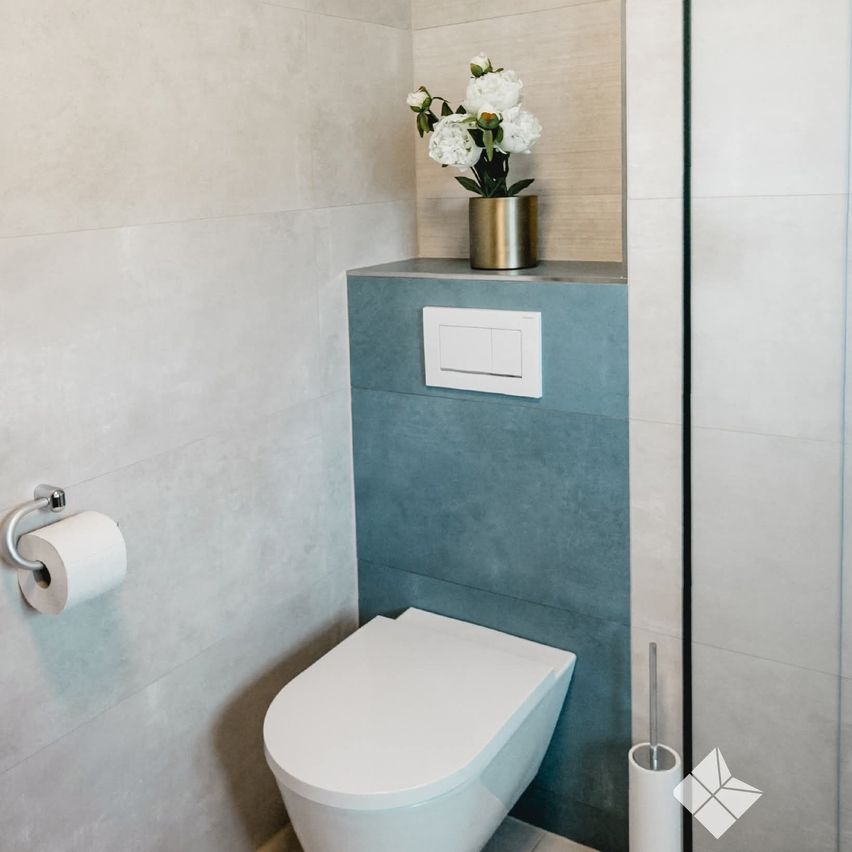 Ein sauberes, modernes Badezimmer mit weißen Wänden und blauen Akzenten. Eine Toilettenschüssel neben einem blauen Regal mit einem weißen Schalter. Ein Blumenvase mit weißen Blumen steht auf dem Regal.