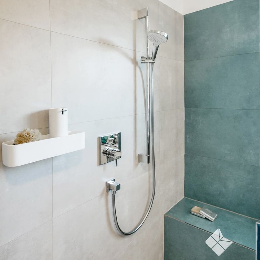 Ein moderner Duschbereich mit einer silbernen Duschbrause und einem weißen Regal. Ein weißes Handtuch und ein Schwamm befinden sich auf dem Regal. Die Dusche befindet sich neben einer weißen und türkisfarbenen gefliesten Wand.