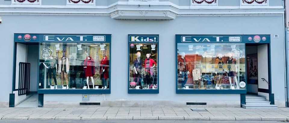 Ein Gebäude mit drei Schaufenstern, in denen jeweils Mannequins mit Kleidung zu sehen sind. Das linke und rechte Schaufenster trägt den Namen Eva T, das mittlere sagt Kinder.