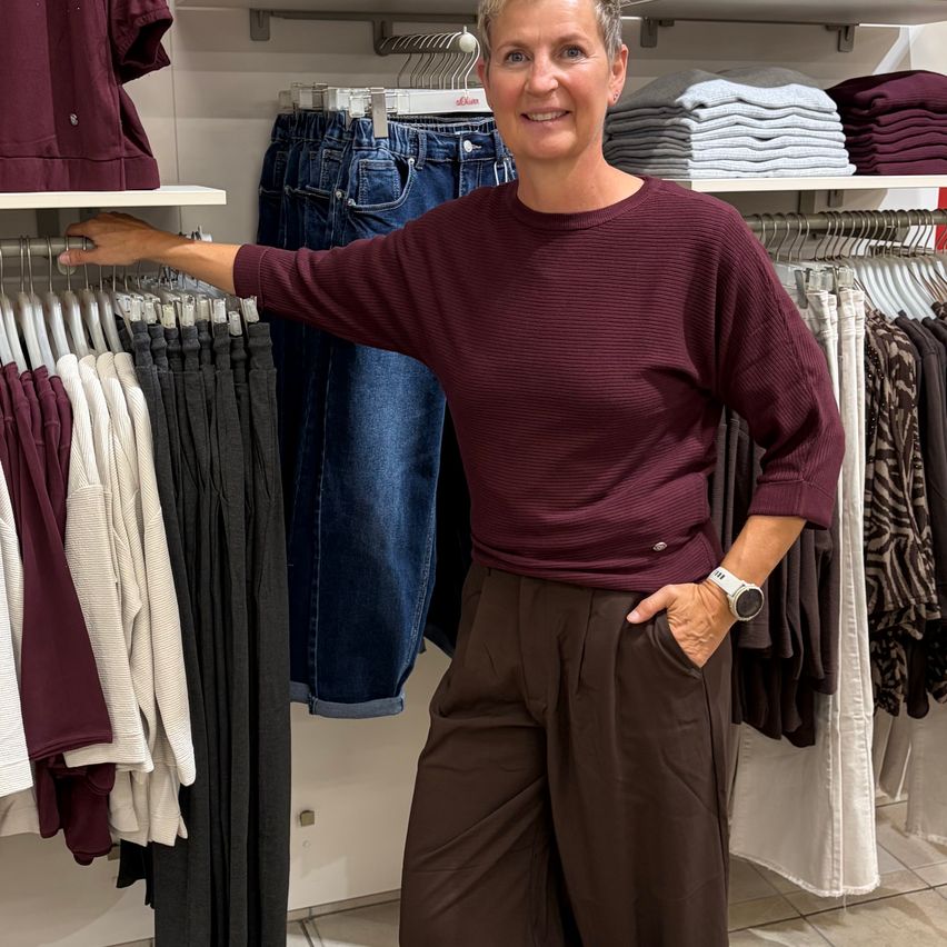A woman in a maroon sweater stands in a clothing store, smiling and posing with her hand in her pocket. Behind her, clothes hang on racks, including jeans and white shirts.