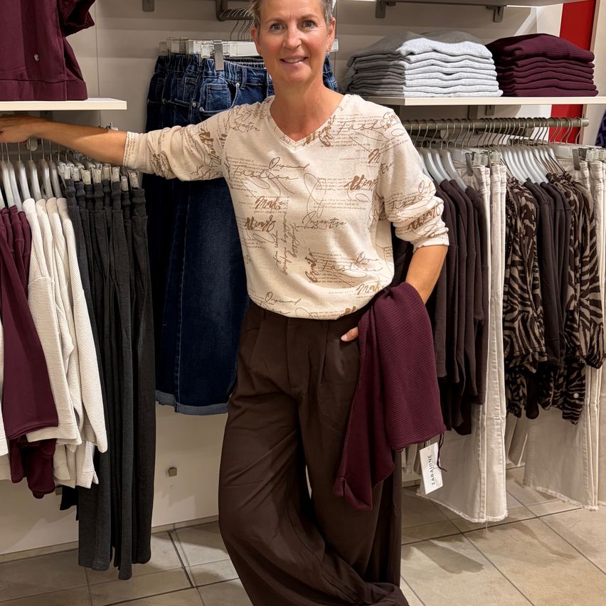 A woman stands in a clothing store, smiling, with her hand on a clothing rack and holding a maroon sweater.