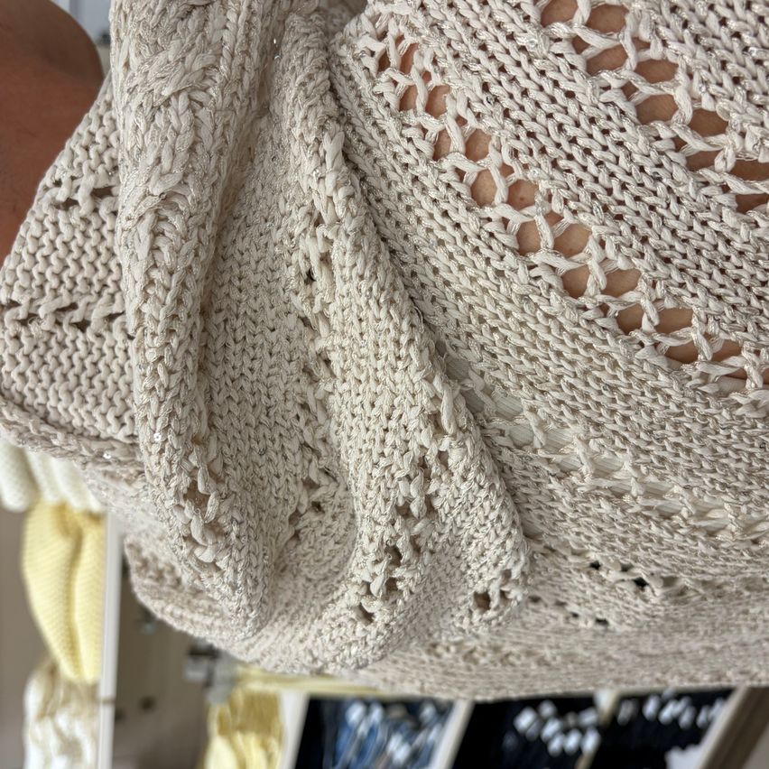 Ein Nahaufnahme eines beigen gestrickten Stoffs mit einem Spitzenmuster. Es ist an einem Kleiderbügel in einem Bekleidungsgeschäft ausgestellt.
