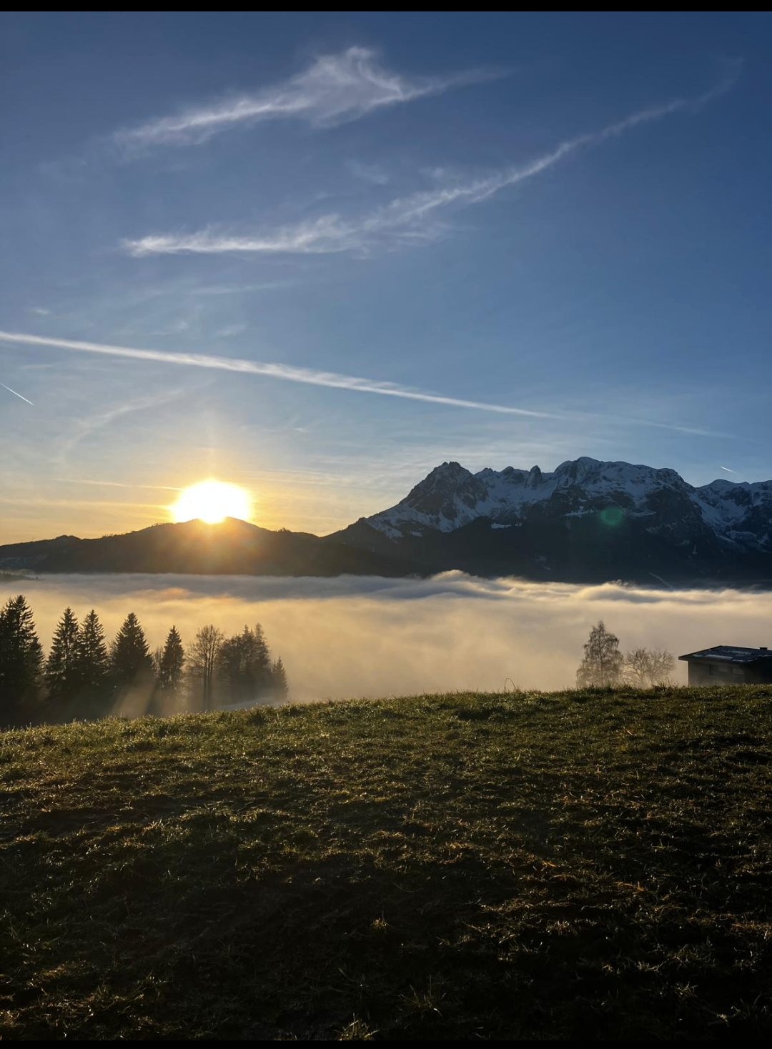 Sonnenuntergang über einer bergigen Landschaft mit einem Vordergrund aus Nebel und Bäumen, wobei ein entferntes Haus zu sehen ist.