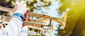Ein Musiker spielt eine goldene Trompete im Freien, mit Bäumen und Sonnenlicht im Hintergrund.