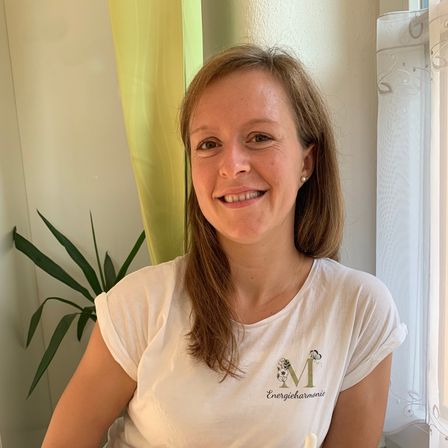 Eine lächelnde Frau in einem weißen T-Shirt hält eine kleine Flasche und sitzt in einem Zimmer mit einer Pflanze und einem Fenster.