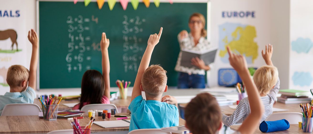 In einem Klassenzimmer heben Kinder die Hände, und eine Lehrerin unterrichtet vorne. Die Tafel hat Matheaufgaben, und eine Karte von Europa ist an der Wand.