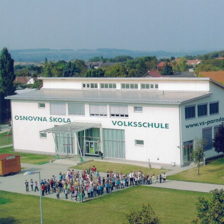 Ein Luftbild einer Schule namens Volksschule mit Solarpaneelen auf dem Dach und einer Menschenmenge vor dem Gebäude.