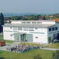 Ein Luftbild einer Schule namens Volksschule mit Solarpaneelen auf dem Dach und einer Menschenmenge vor dem Gebäude.