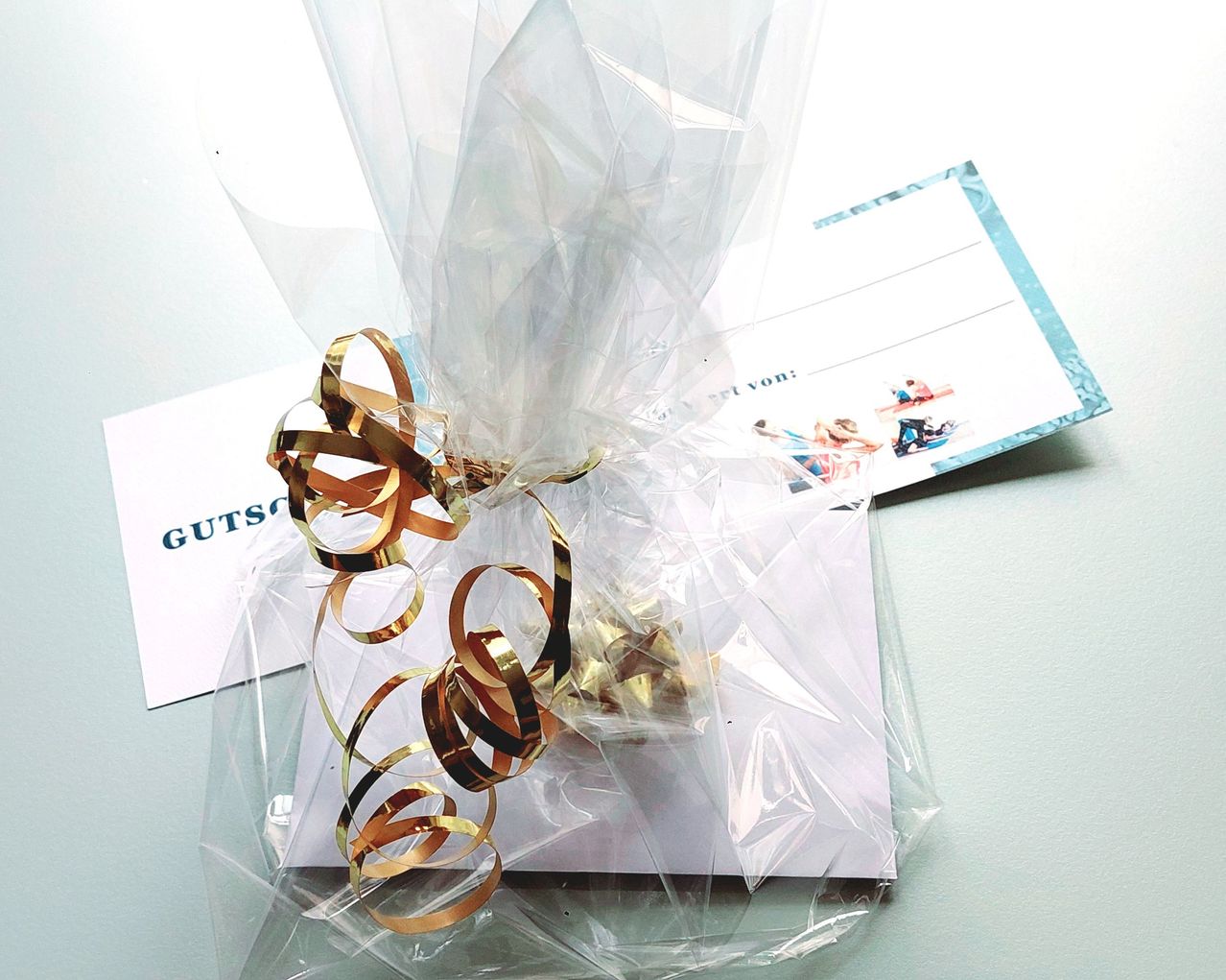Eine durchsichtige Plastiktüte mit goldenen Bändern enthält ein verpacktes Geschenk. Die Tüte liegt auf einer weißen Karte mit blauen Rändern und dem Wort 'Gutschein'. Hinter der Tüte ist ein blauer Umschlag zu sehen.