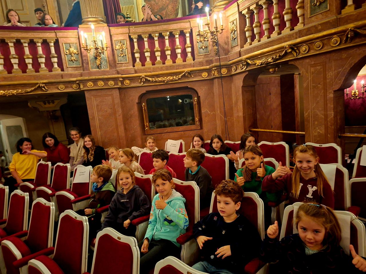 Eine Gruppe von Kindern sitzt in roten Stühlen in einem Theater und gibt Daumen hoch. Das Theater hat goldene Dekorationen und Kronleuchter.
