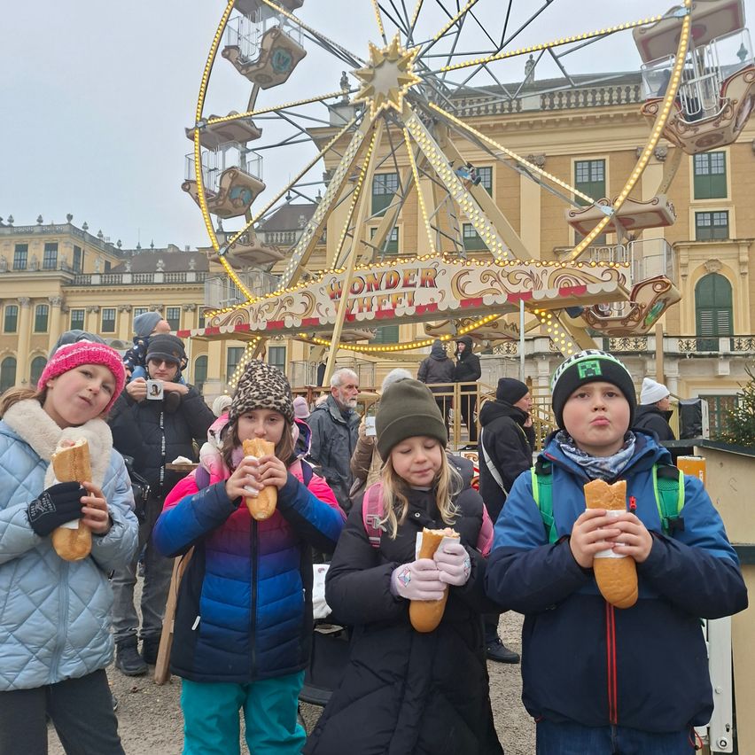 Vier Kinder essen Brot vor einem Riesenrad, das vor einem großen Gebäude mit vielen Fenstern steht.