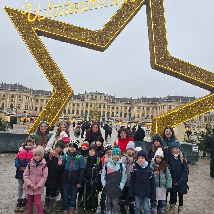 Eine Gruppe von Kindern und zwei Erwachsenen posiert für ein Foto vor einer goldenen sternförmigen Struktur, mit einem großen Gebäude und Weihnachtsdekorationen im Hintergrund.