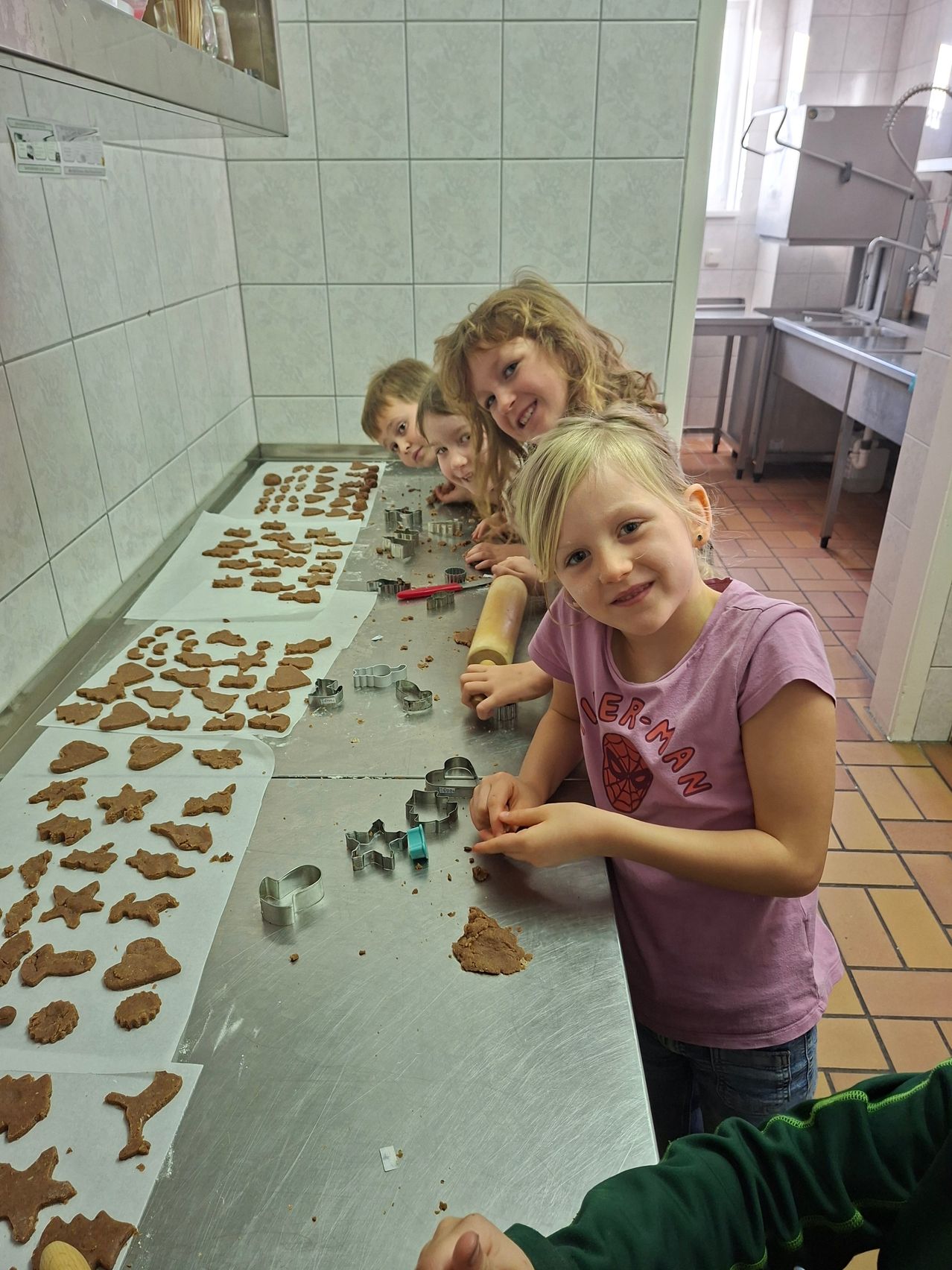 Vier Kinder, eines in einem rosa Shirt, backen Kekse an einem Tisch mit Ausstechern und Teig.