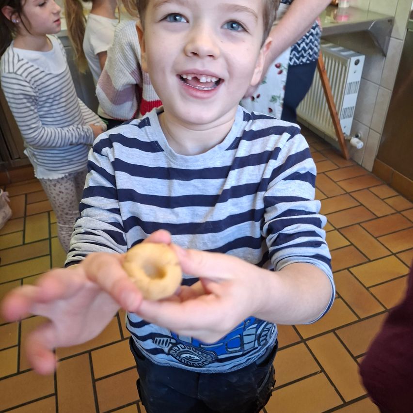 Ein junger Junge hält einen Donut in einem Raum mit anderen Kindern, einem weißen Heizkörper und gefliestem Boden.