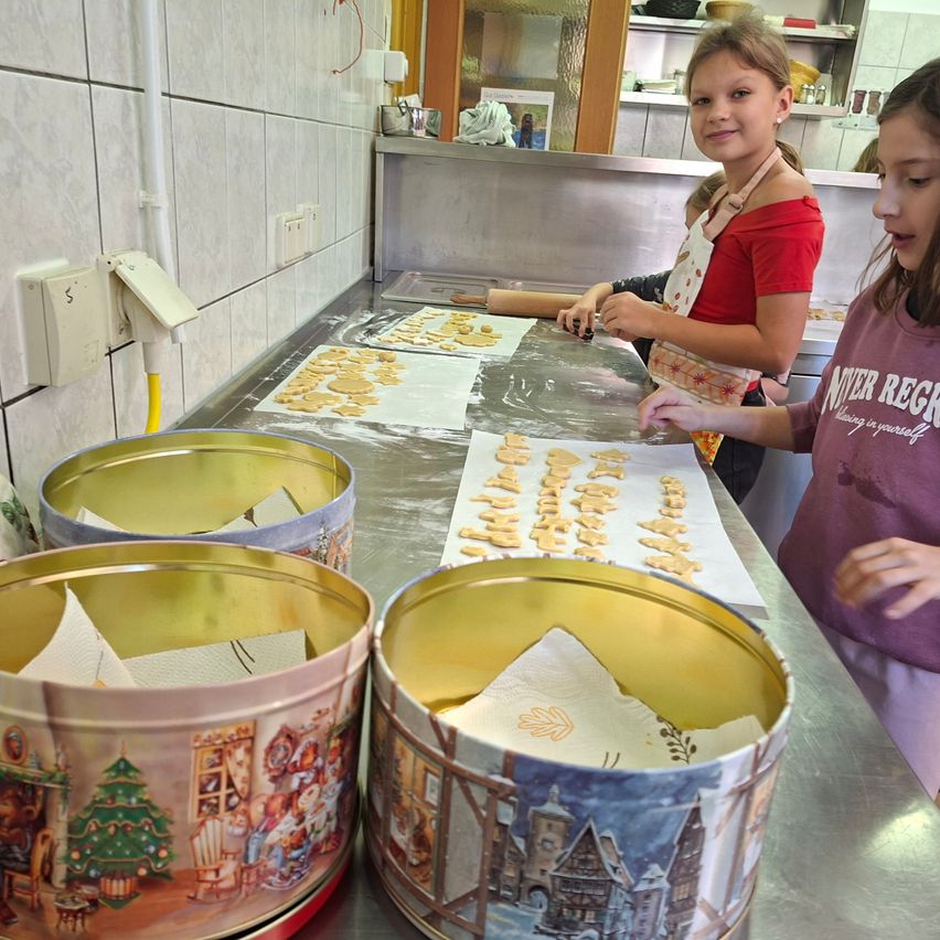 Zwei Mädchen sind in einer Küche und bereiten Kekse zu. Ein Mädchen benutzt einen Nudelholz auf einem Tablett, während das andere Mädchen Kekse auf einem anderen Tablett handhabt. Vor ihnen stehen zwei große Blechdosen mit Papiertüchern darin.