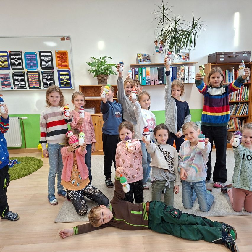 Eine Gruppe von Kindern in einem Klassenzimmer, posiert für ein Foto. Sie alle halten selbstgemachte Schneemann-Spielzeuge und lächeln fröhlich. Der Raum hat Regale mit Büchern und Pflanzen.