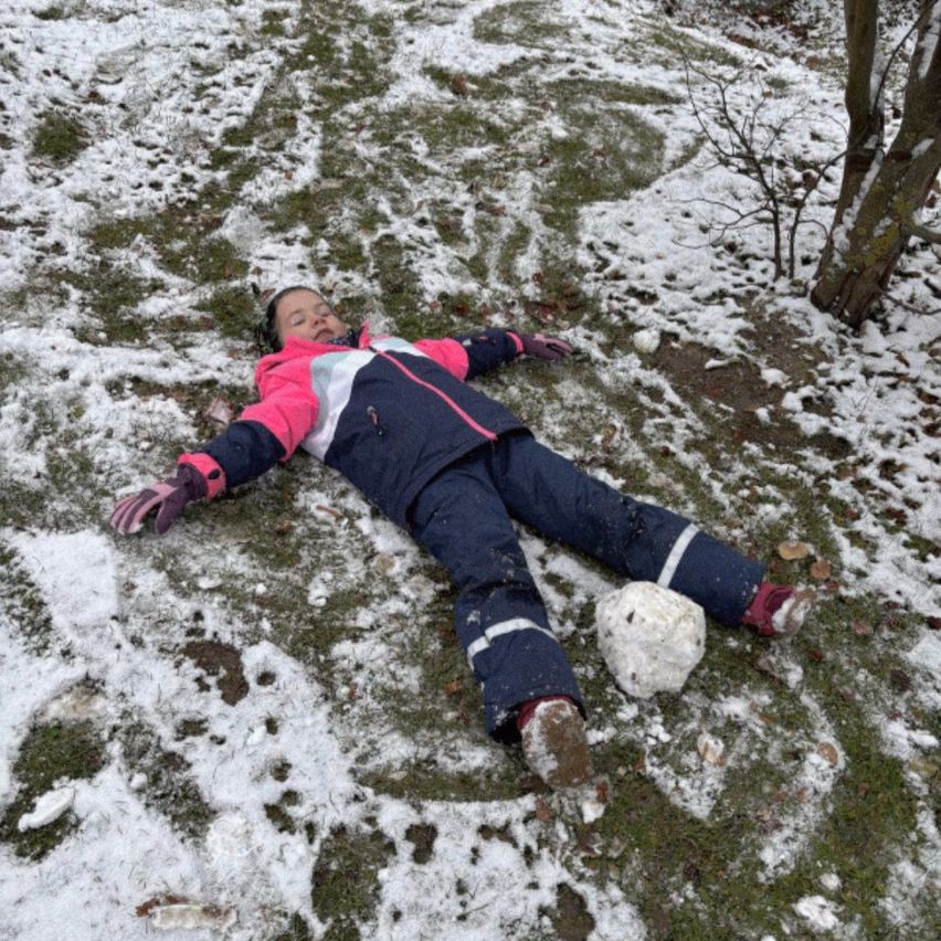 Ein junges Mädchen liegt auf dem Rücken im Schnee, die Arme weit ausgestreckt, mit einem Schneemann in der Nähe. Sie trägt eine rosa-blaue Winterjacke.