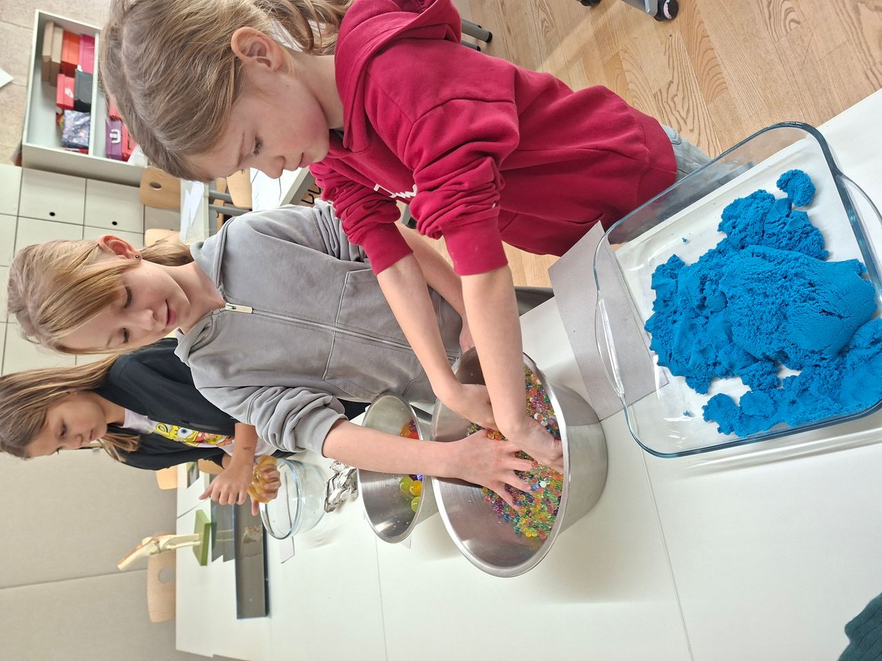 Ein junges Mädchen in einer roten Kapuzenjacke schüttet bunte Streusel in eine Schüssel. Eine weitere junge Frau in Grau ist in der Nähe und beteiligt sich ebenfalls an der Aktivität.
