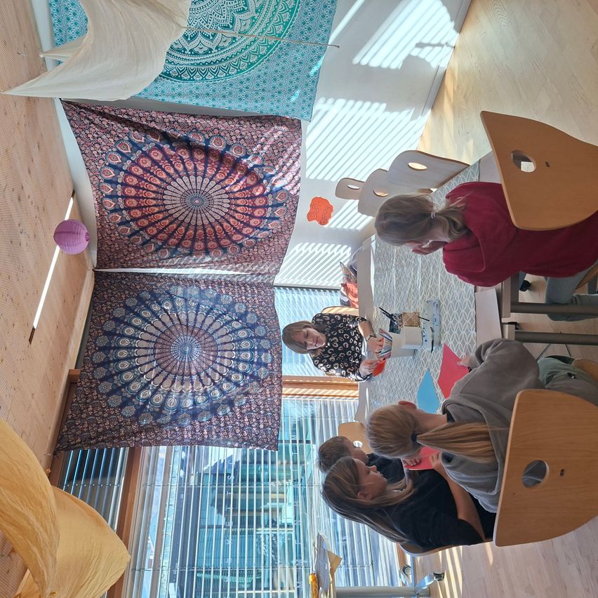 Eine obere Ansicht eines Raums mit gemusterten Wandteppichen. Eine Frau steht in der Mitte, und mehrere Frauen sitzen um einen Tisch. Dekorative Gegenstände und Lampen hängen von der Decke.