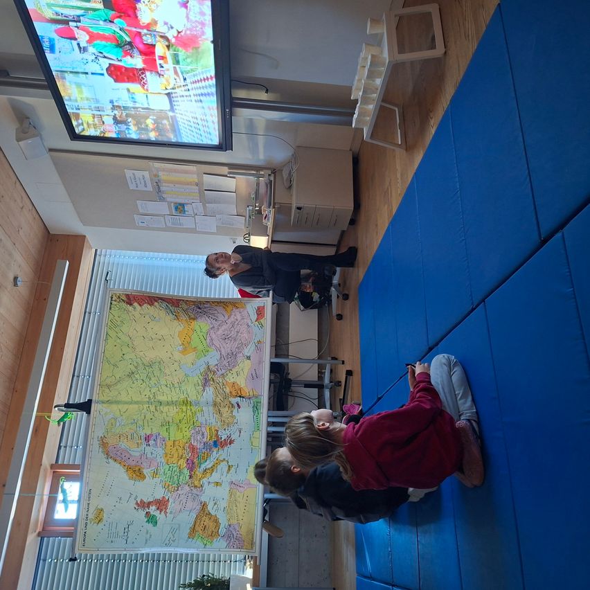 Eine Frau steht vorne in einem Klassenzimmer und unterrichtet eine Gruppe von Kindern, die auf blauen Matten sitzen. Eine große Karte hängt an der Wand hinter ihnen.