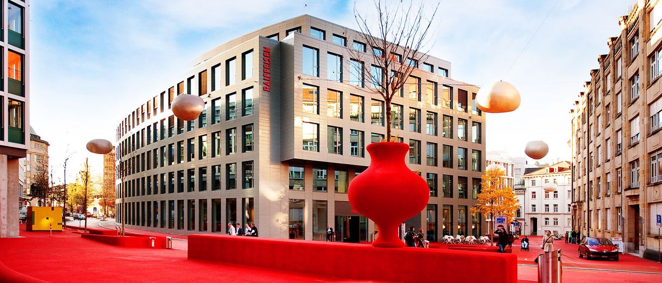 Ein modernes Gebäude mit Glasfenstern und dem Wort 'Raiffeisen' an der Seite. Ein roter Vasen mit einem kleinen Baum steht vor dem Gebäude. Ein roter Teppich liegt vor dem Gebäude, auf dem Menschen spazieren.