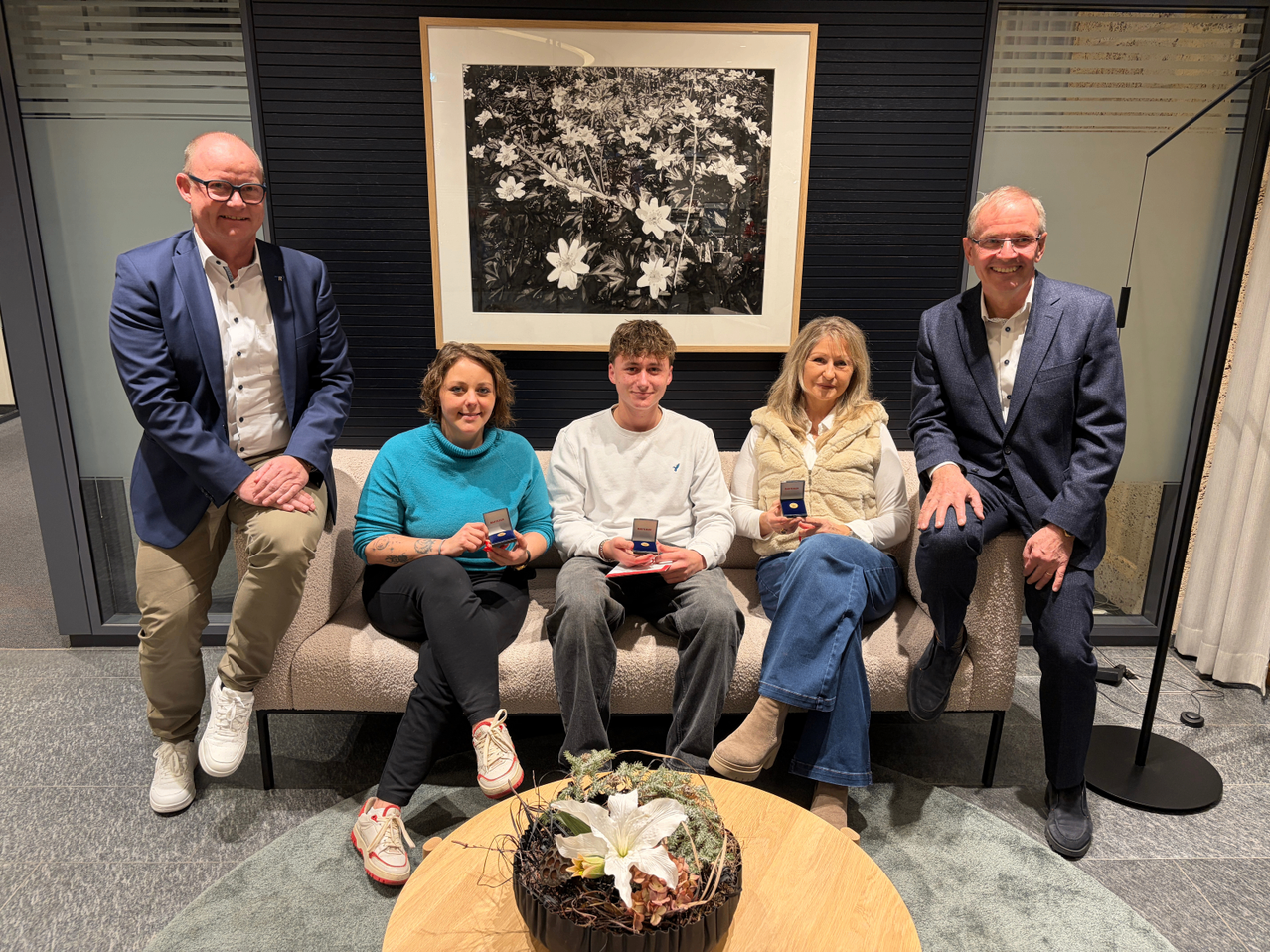Fünf Personen sitzen auf einer Couch und lächeln für ein Foto. Ein Mann und eine Frau an den Enden halten Auszeichnungen. Ein gerahmtes Foto mit weißen Blumen hängt an der Wand hinter ihnen.