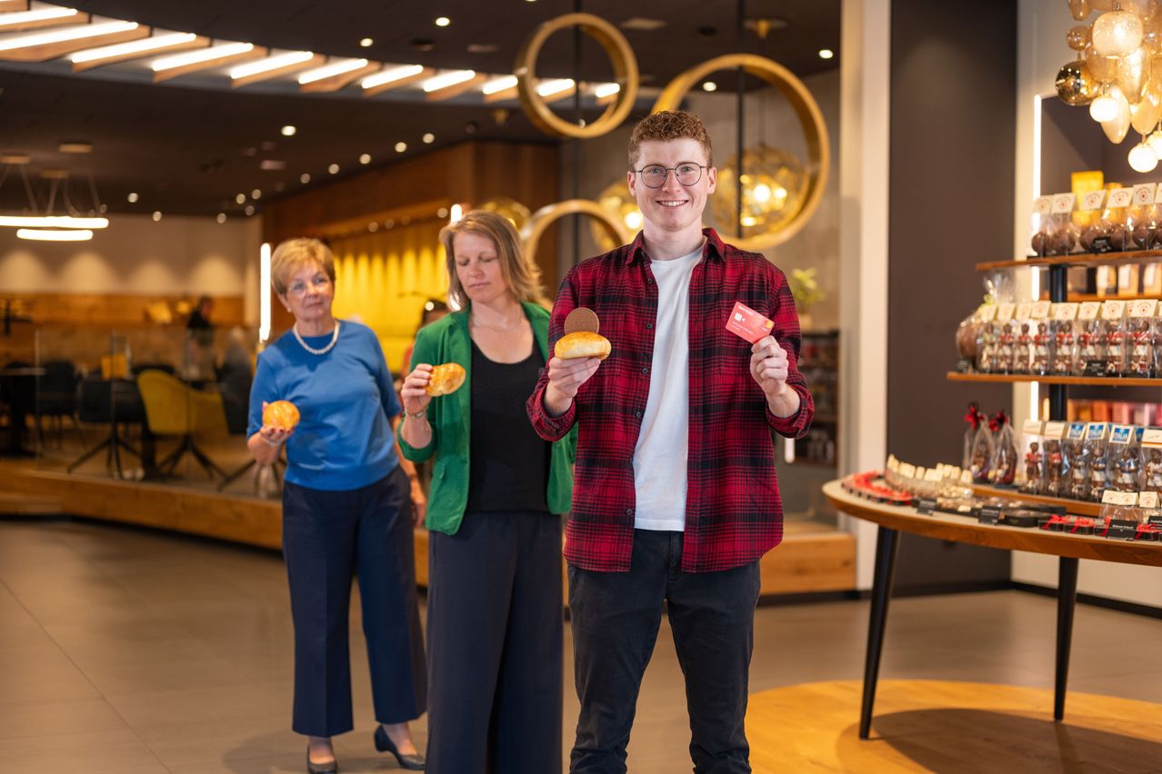 Three people stand in a shop. A man in a red plaid shirt holds cookies and a card. Two women, one in blue and one in green, also hold cookies.