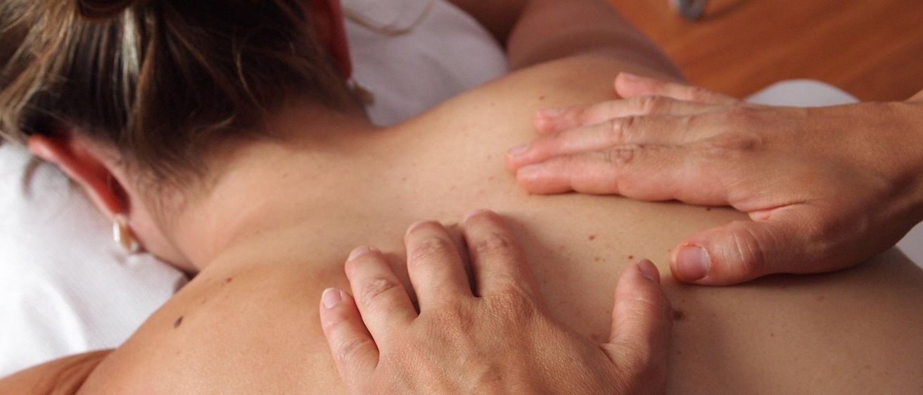 Eine Frau liegt auf einem Bett und erhält eine Massage auf ihrem oberen Rücken von einer Person, die Hände auf ihren Schultern und Rücken hat.