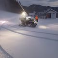 Zwei Arbeiter räumen Schnee von einem Hang mit einem Fahrzeug, das oben ein Licht hat. Ein kleines Gebäude ist im Hintergrund zu sehen.