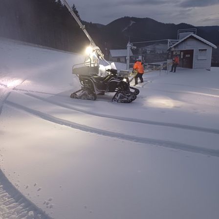 Zwei Arbeiter räumen Schnee von einem Hang mit einem Fahrzeug, das oben ein Licht hat. Ein kleines Gebäude ist im Hintergrund zu sehen.
