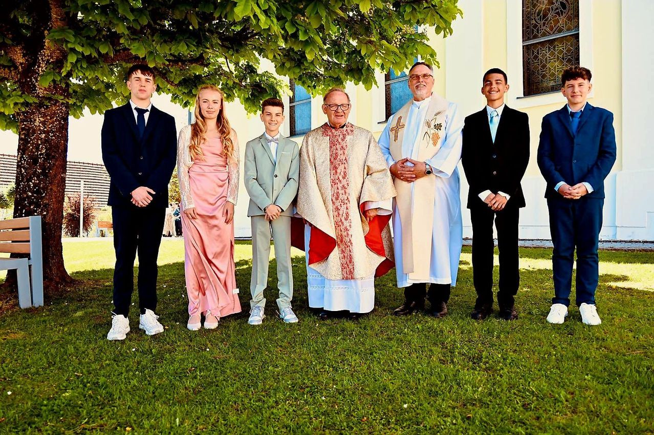 Mehrere Menschen stehen draußen, wahrscheinlich bei einer Kirchenveranstaltung. Sie sind formell gekleidet, mit zwei Priestern in der Mitte. Die beiden jungen Leute vorne lächeln.