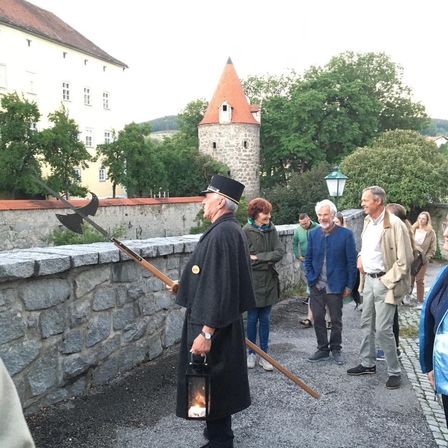 Ein älterer Mann in einem schwarzen Gewand hält eine Laterne und eine Lanze und führt eine Gruppe von Menschen auf einem Kiesweg in der Nähe einer Steinmauer und eines Schlosses.