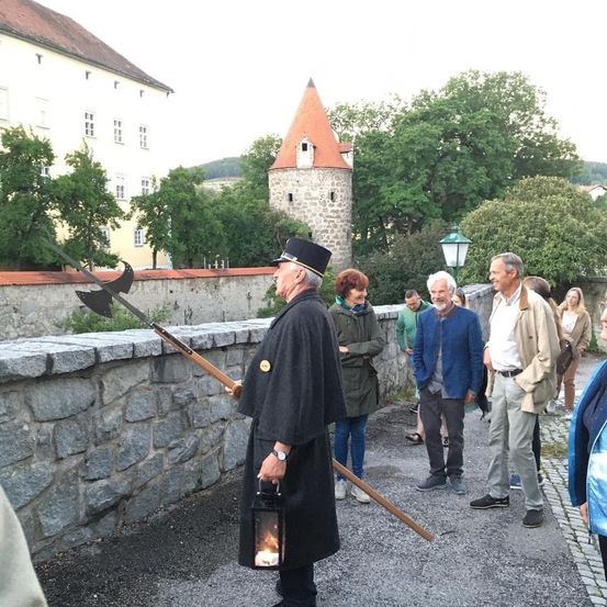 Ein älterer Mann in einem schwarzen Gewand hält eine Laterne und eine Lanze und führt eine Gruppe von Menschen auf einem Kiesweg in der Nähe einer Steinmauer und eines Schlosses.