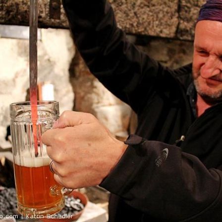 Ein Mann in einer schwarzen Jacke schenkt Bier in einen Glaskrug in einer rustikalen Umgebung.