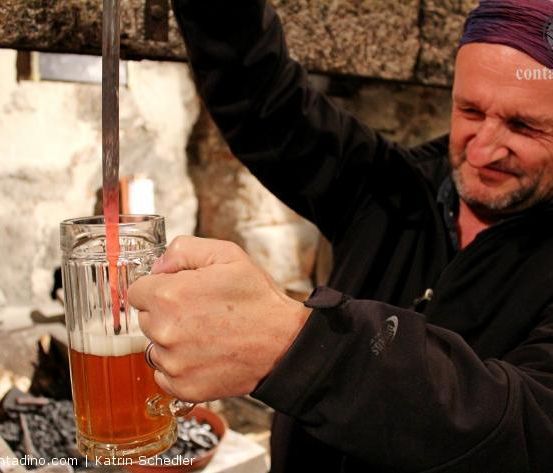 Ein Mann in einer schwarzen Jacke schenkt Bier in einen Glaskrug in einer rustikalen Umgebung.
