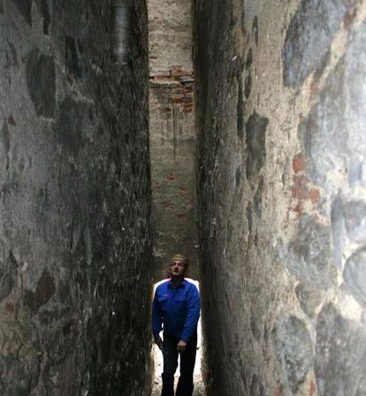 Ein Mann steht in einer engen Gasse zwischen zwei Steinmauern. Die Mauern sind alt und verwittert. Er trägt eine blaue Jacke und schwarze Hosen.