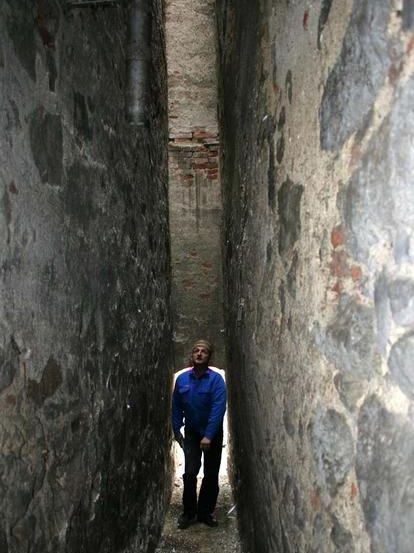 Ein Mann steht in einer engen Gasse zwischen zwei Steinmauern. Die Mauern sind alt und verwittert. Er trägt eine blaue Jacke und schwarze Hosen.