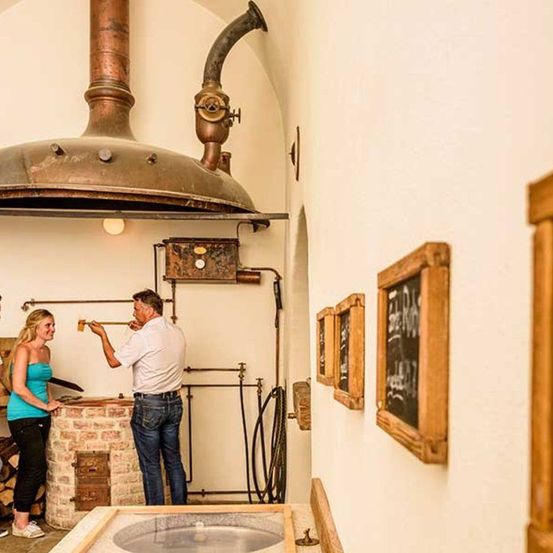 Ein Mann und eine Frau stehen in einem Raum mit einem großen Kupfertopf, Rohren und einem Ziegelofen. Der Mann hält einen Holzstab, möglicherweise zum Kochen. An der Wand hängen gerahmte Tafeln mit Schrift.