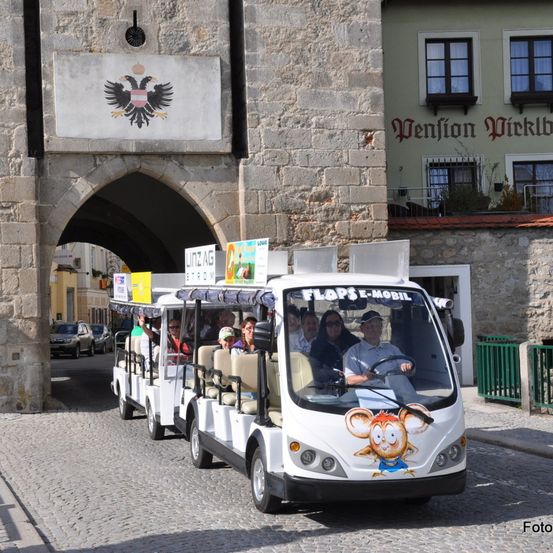 Ein weißer Trolley mit einem Cartoon-Mäuschen auf der Vorderseite fährt durch einen historischen Torbogen. Menschen sitzen auf Bänken. Ein Schild steht für Linz AG.