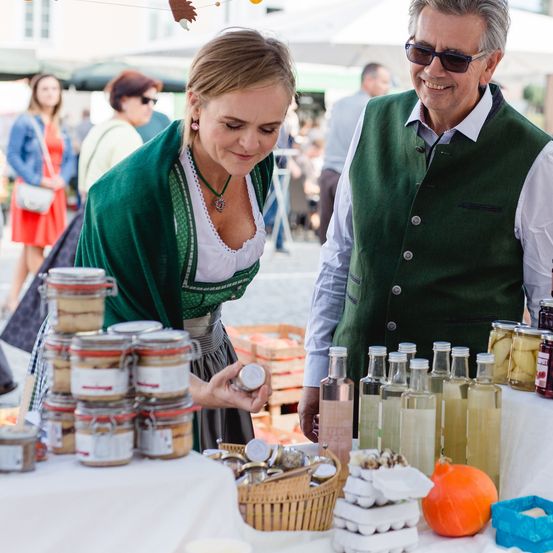 Eine Frau in einem grünen Outfit und ein Mann in einer Weste stehen an einem Marktstand, untersuchen Gläser mit Lebensmitteln und Flaschen mit Öl. Hinter ihnen gehen mehrere Leute herum, und es gibt Zelte und Gebäude im Hintergrund.
