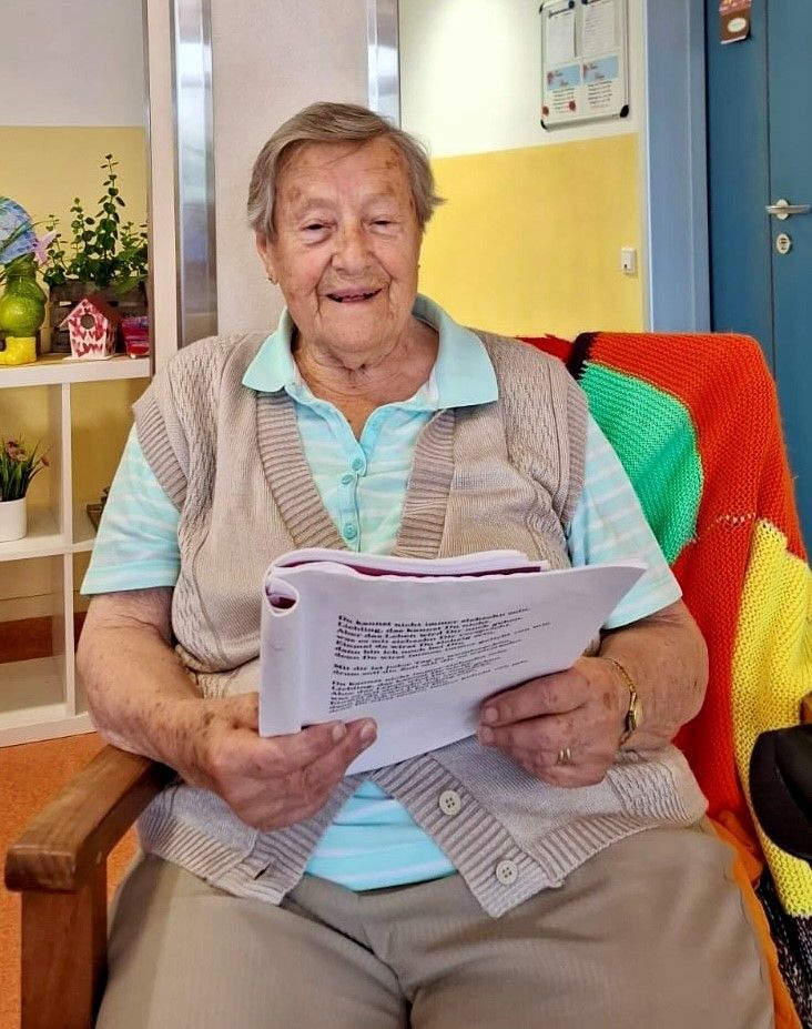 Eine ältere Frau sitzt auf einem Stuhl und hält ein Dokument mit Text in der Hand, lächelt und hat eine gestrickte Decke auf dem Schoß. Hinter ihr ist eine farbenfrohe Wand mit Regalen und Pflanzen.
