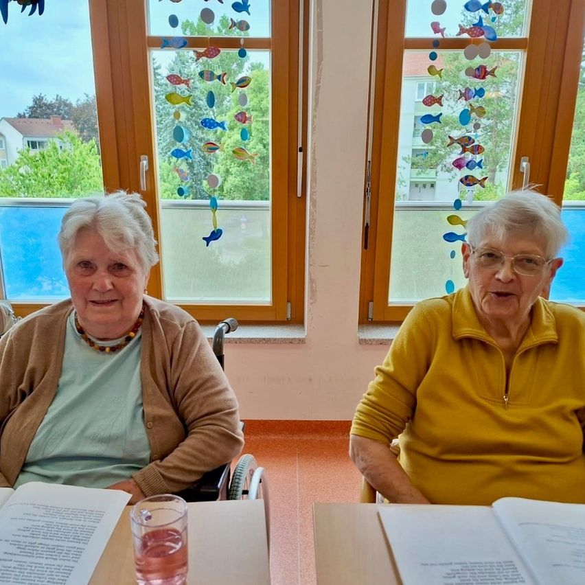 Zwei ältere Frauen sitzen an einem Tisch, eine im Rollstuhl, beide lächeln, mit Büchern und einem Glas mit rosa Flüssigkeit vor ihnen. Hinter ihnen befinden sich zwei Fenster mit bunten Fischmobiles. Draußen vor den Fenstern sind Bäume und ein Gebäude.