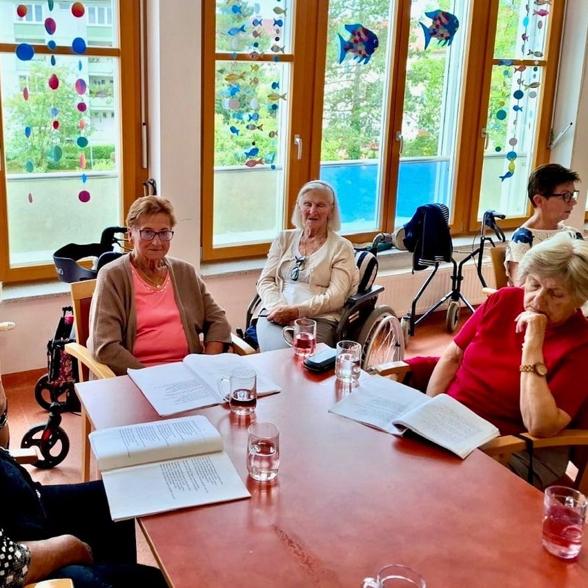 Ältere Frauen sitzen um einen Tisch mit Büchern und Gläsern, zwei davon in Rollstühlen. Sie befinden sich in einem Raum mit großen Fenstern und bunten Fischdekorationen.