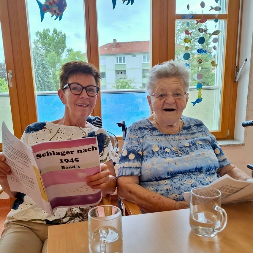 Zwei ältere Frauen sitzen in Rollstühlen und lächeln. Eine liest ein Buch mit dem Titel 'Schlager nach 1945 Band 3'. Beide tragen Brillen und eine trägt eine Halskette. Sie sitzen an einem Fenster mit Fischdekorationen.