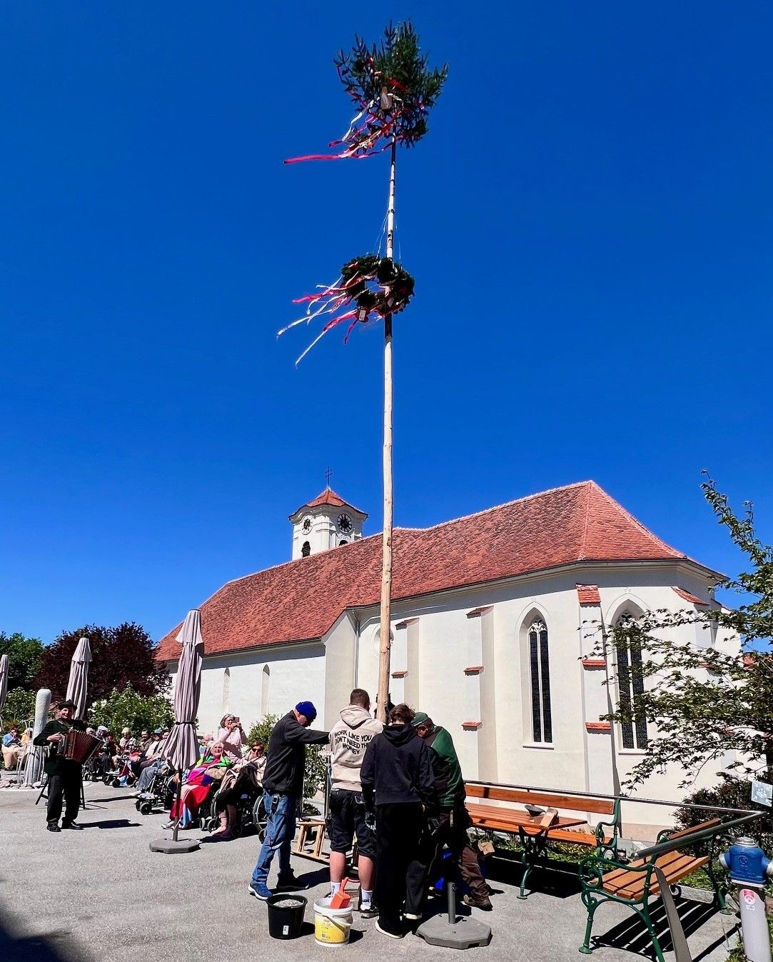 Menschen versammeln sich vor einer Kirche, ein Mann spielt Akkordeon. Ein hoher Pfahl mit Dekorationen steht in der Mitte. Ein Turm und eine Uhr schmücken die Kirche. Bäume und Bänke sind im Vordergrund.
