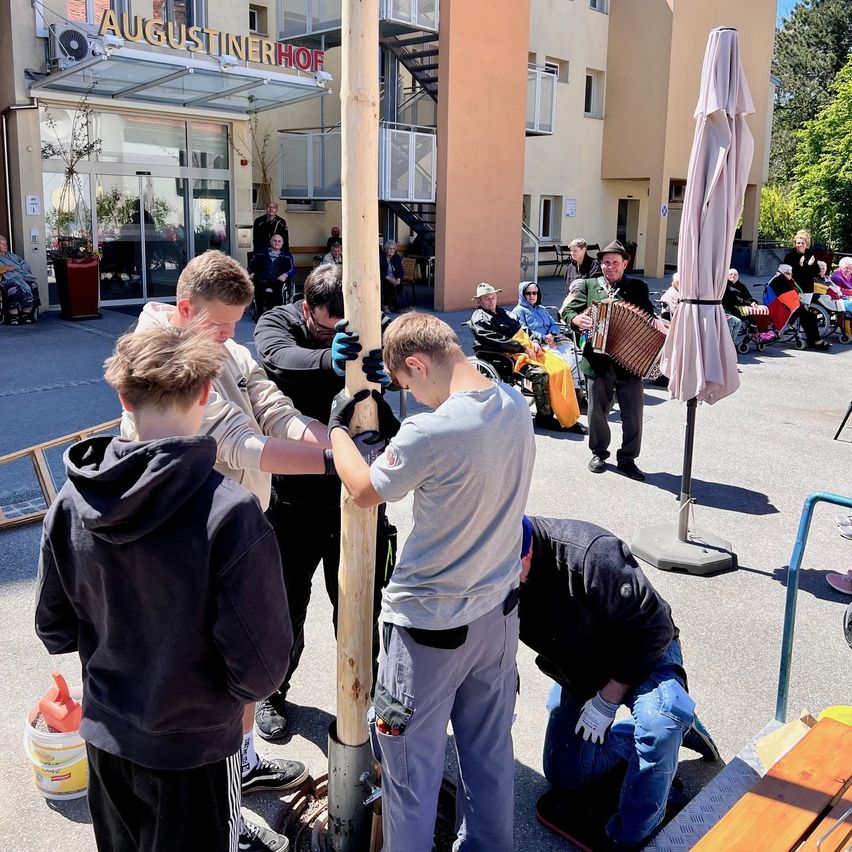 Mehrere Menschen sind im Freien, möglicherweise bei einer Flaggenzeremonie. Sie stehen um einen hohen Pfahl herum, und einer hält ein Akkordeon. Hinter ihnen ist ein Gebäude mit dem Namen 'AUGUSTINIERHOF' zu sehen.