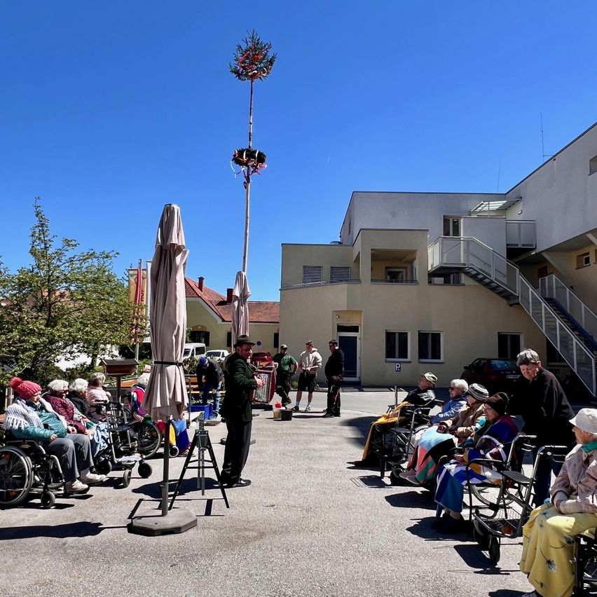 Ein Mann steht neben einem Baum mit Dekorationen, während ältere Menschen in Rollstühlen unter Schirmen auf einem Parkplatz sitzen. Dahinter steht ein Gebäude mit Treppen und einem geparkten Auto.