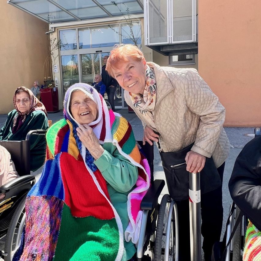 Eine ältere Frau im Rollstuhl lächelt für ein Foto, bedeckt mit einer bunten Decke, neben ihr steht eine andere Frau am Eingang eines Gebäudes.